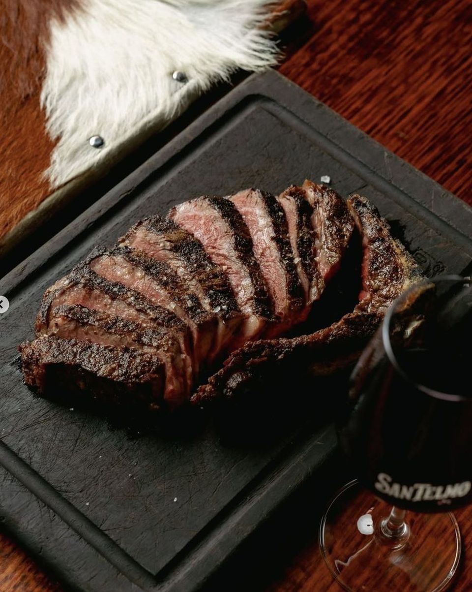 A sliced, grilled steak on a black board beside a San Telmo wine glass